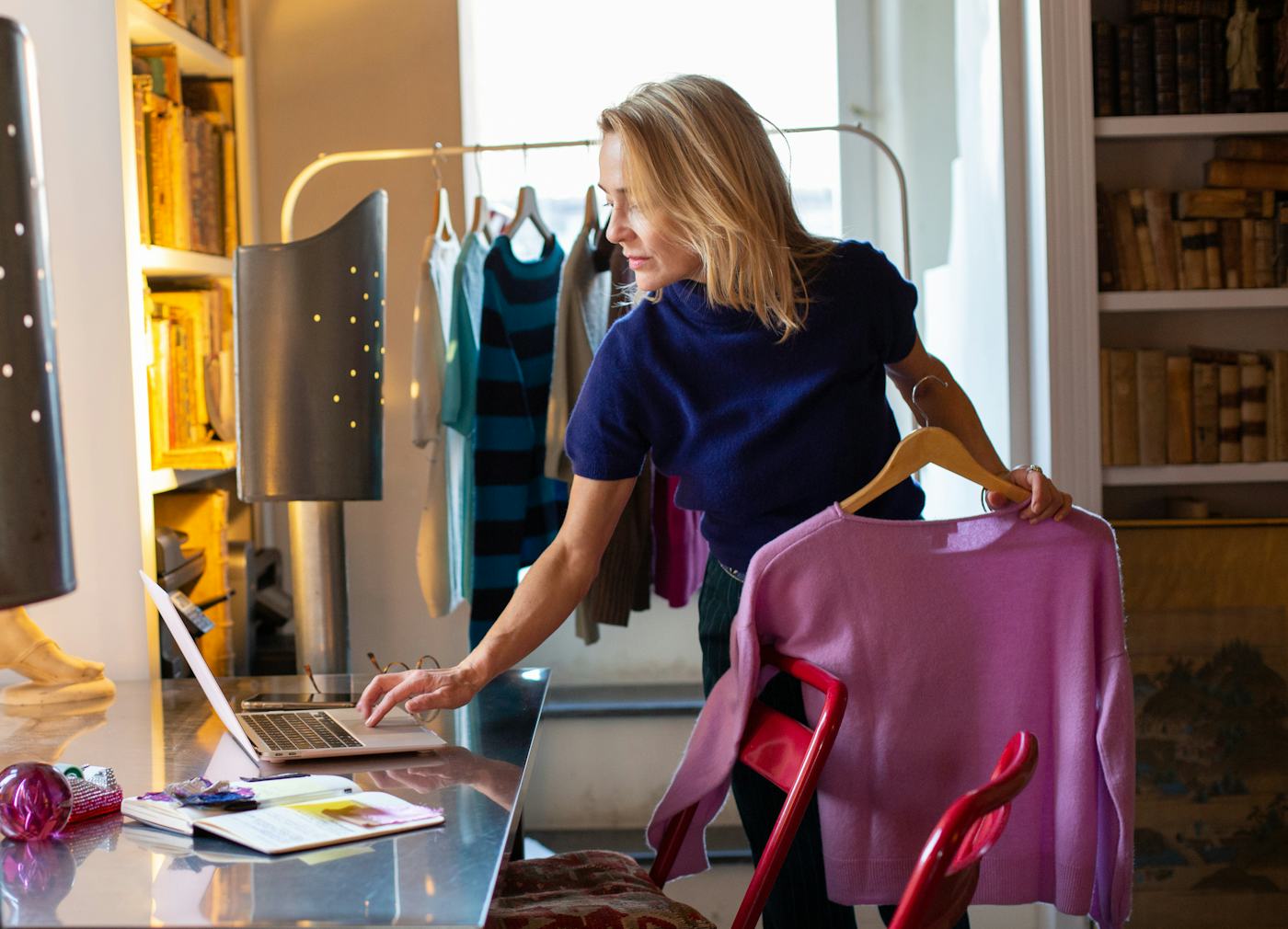 A woman in a navy top stands at a desk, checking a laptop while holding a pink sweater. Colorful clothing hangs in the background, creating a vibrant workspace.