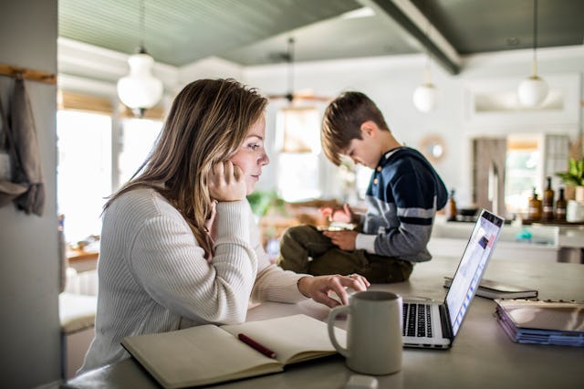 A woman sits at a kitchen counter working on a laptop while a young boy stands nearby, focused on a ...