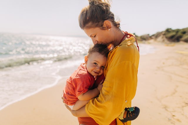 A woman in a yellow blouse joyfully hugs a smiling child in a red shirt, standing on a sunny beach w...