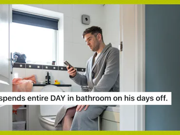 Man in a bathrobe sits on a toilet, looking at his phone. The bathroom has white tiles and a window ...
