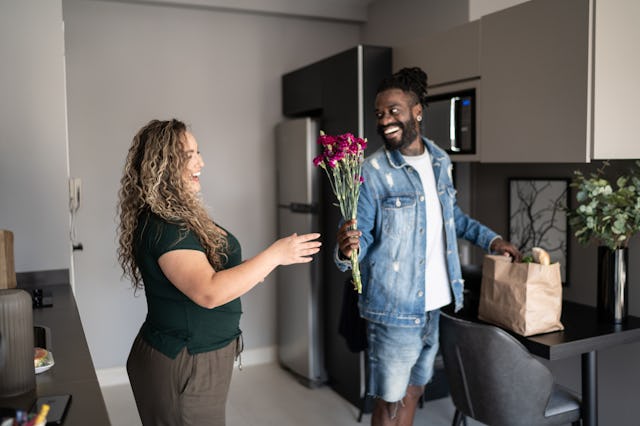 A man in a denim jacket gives flowers to a smiling woman in a kitchen. A grocery bag is on the table...