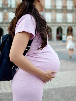 Pregnant woman in a pink dress with a black backpack stands in a plaza, hands on belly. Blurred peop...