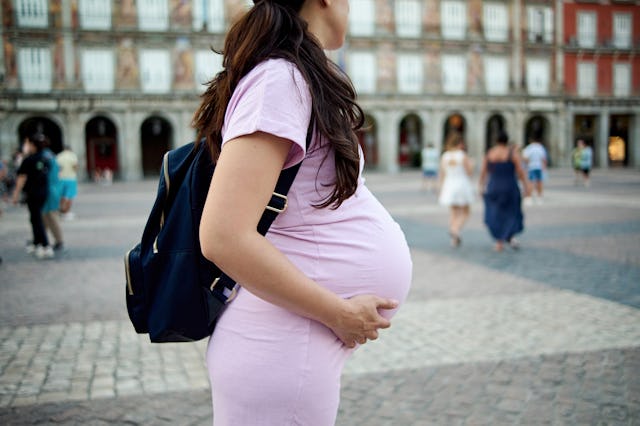 Pregnant woman in a pink dress with a black backpack stands in a plaza, hands on belly. Blurred peop...