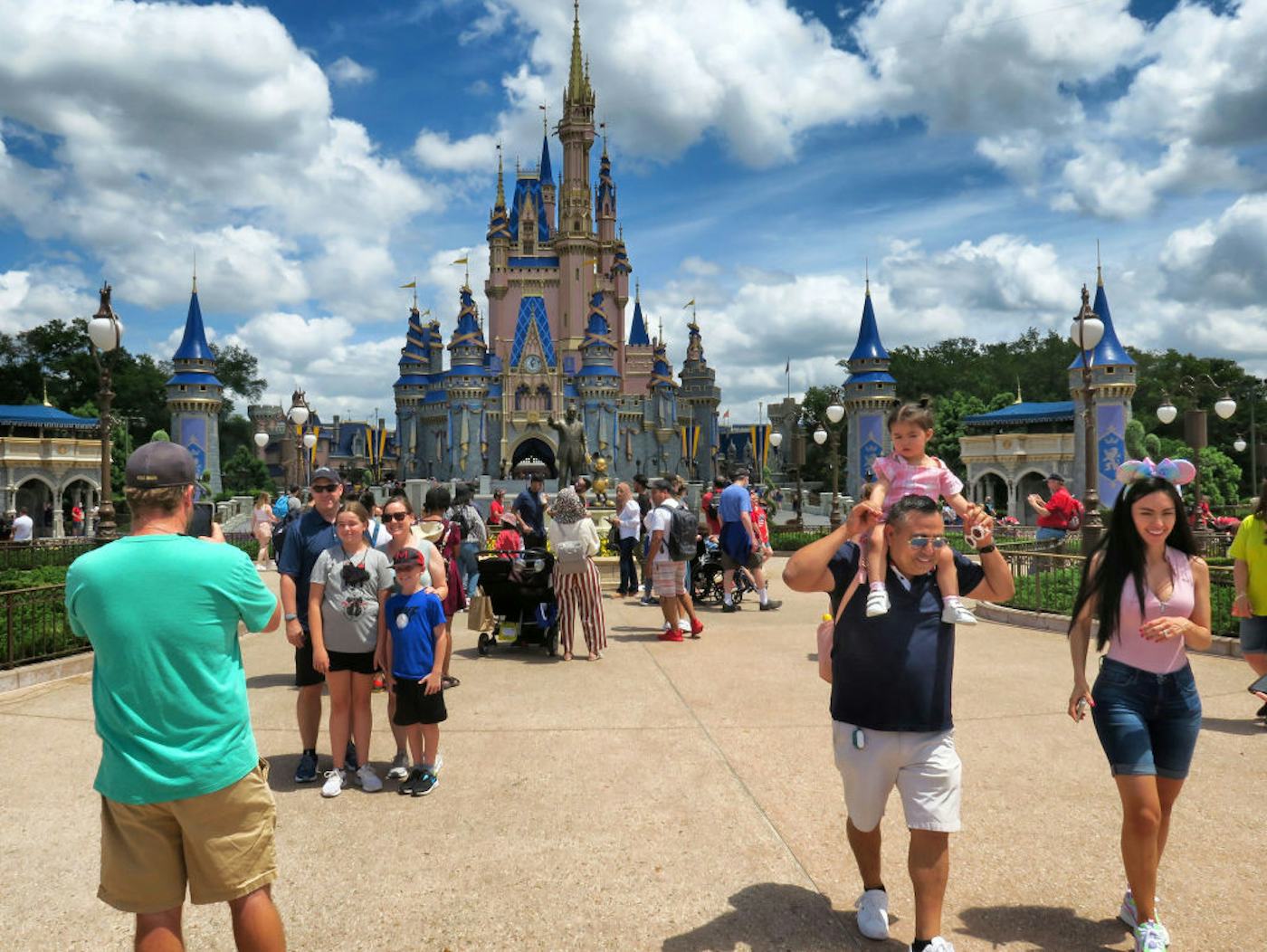 A crowd of people at a theme park, with a large castle in the background. Visitors are taking photos and walking under a partly cloudy sky.
