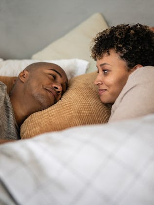 A couple lies in bed, wrapped in a blanket, facing each other and smiling. The room is softly lit, w...