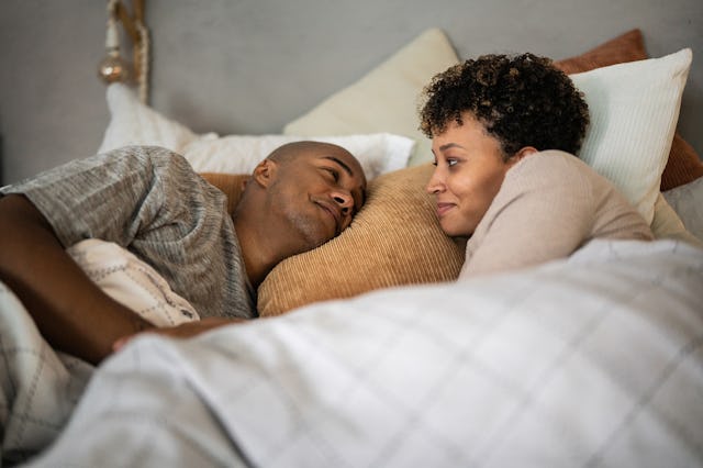 A couple lies in bed, wrapped in a blanket, facing each other and smiling. The room is softly lit, w...