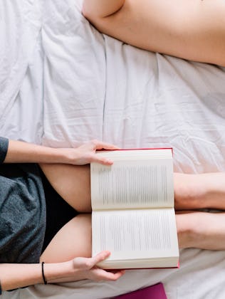 Two people lying on a bed. One, wearing a dark top and black shorts, holds an open book. The other i...
