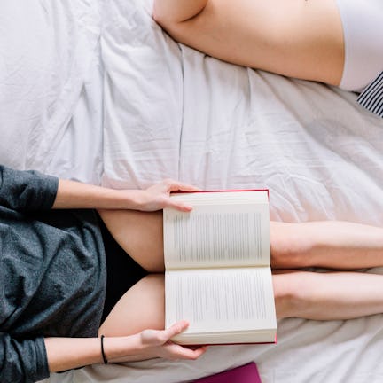 Two people lying on a bed. One, wearing a dark top and black shorts, holds an open book. The other i...