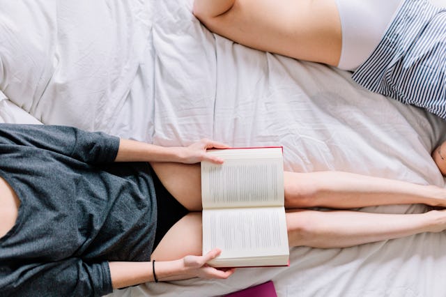 Two people lying on a bed. One, wearing a dark top and black shorts, holds an open book. The other i...