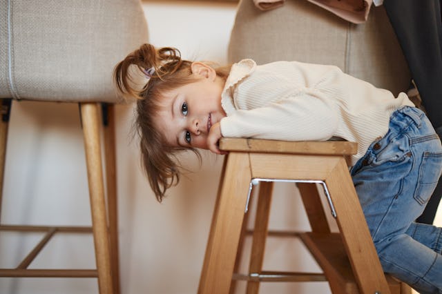 Child with brown hair in a white sweater and blue jeans lying face down on a wooden chair, looking t...