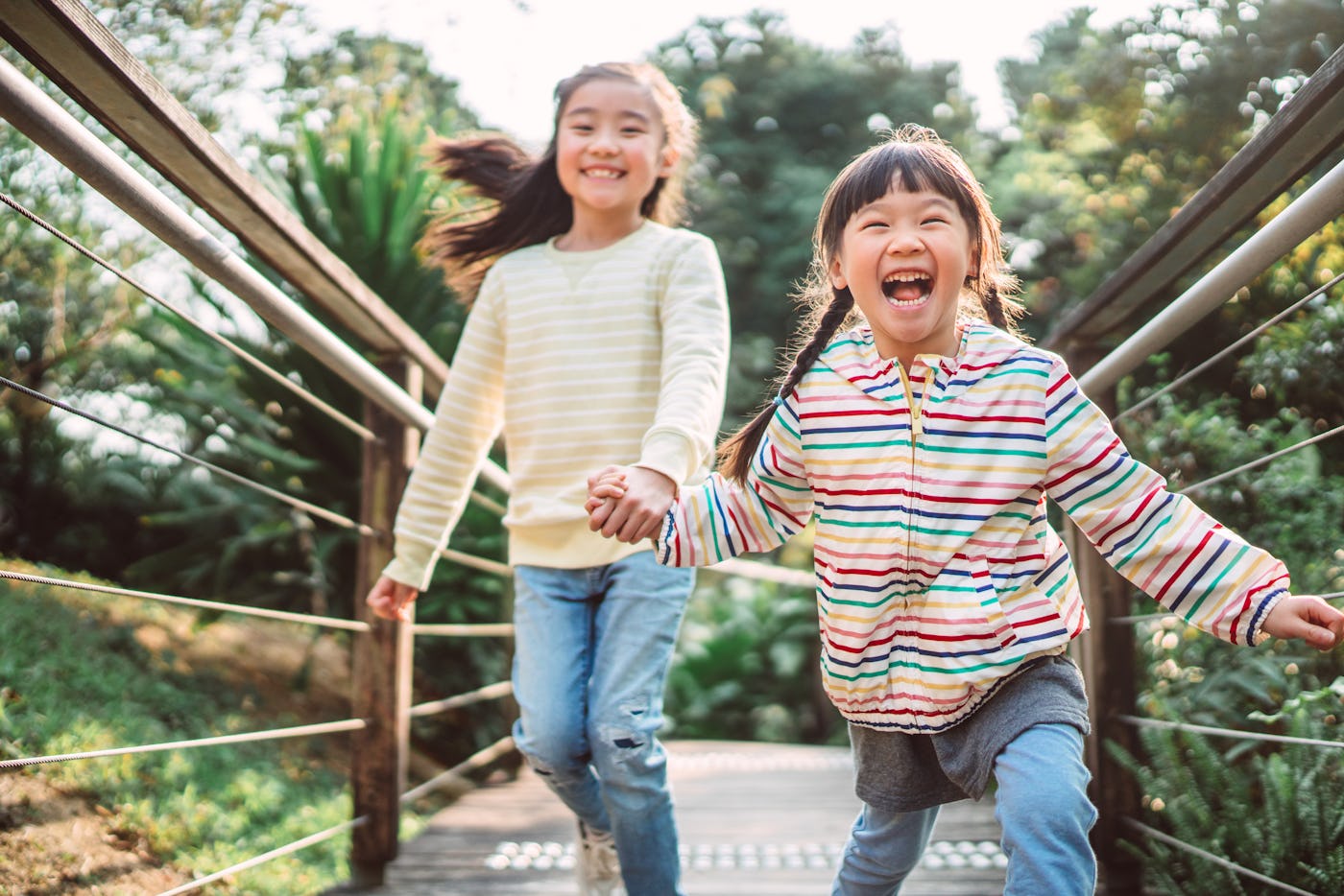 Two children, one in a striped jacket and the other in a striped sweater, joyfully run hand in hand on a wooden bridge surrounded by greenery.