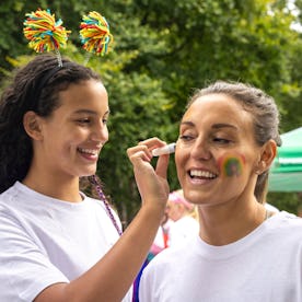 Two women in white t-shirts, one painting a rainbow on the other's face. They smile outdoors near a ...