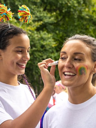 Two women in white t-shirts, one painting a rainbow on the other's face. They smile outdoors near a ...