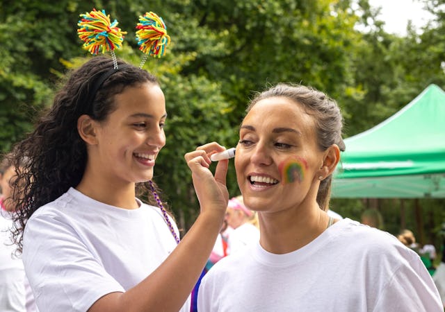 Two women in white t-shirts, one painting a rainbow on the other's face. They smile outdoors near a ...
