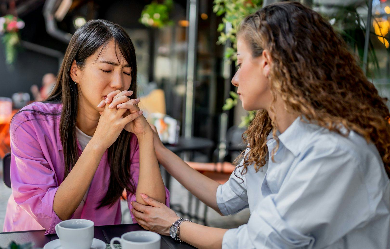 Two women seated at a table outdoors, one comforting the other. The woman in pink appears upset, hol...