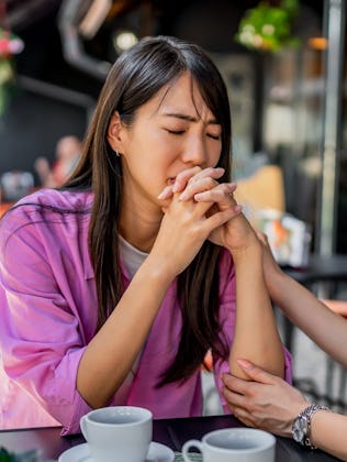 Two women seated at a table outdoors, one comforting the other. The woman in pink appears upset, hol...