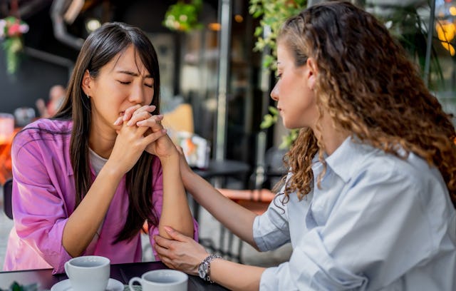 Two women seated at a table outdoors, one comforting the other. The woman in pink appears upset, hol...
