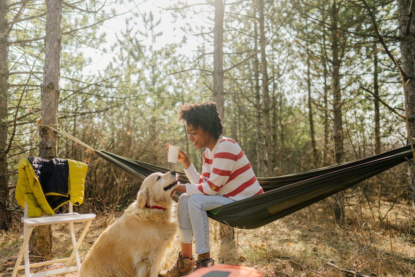 Person sitting in a hammock in a forest, holding a mug, petting a golden retriever. A jacket hangs on a nearby chair. Sunlight filters through trees.