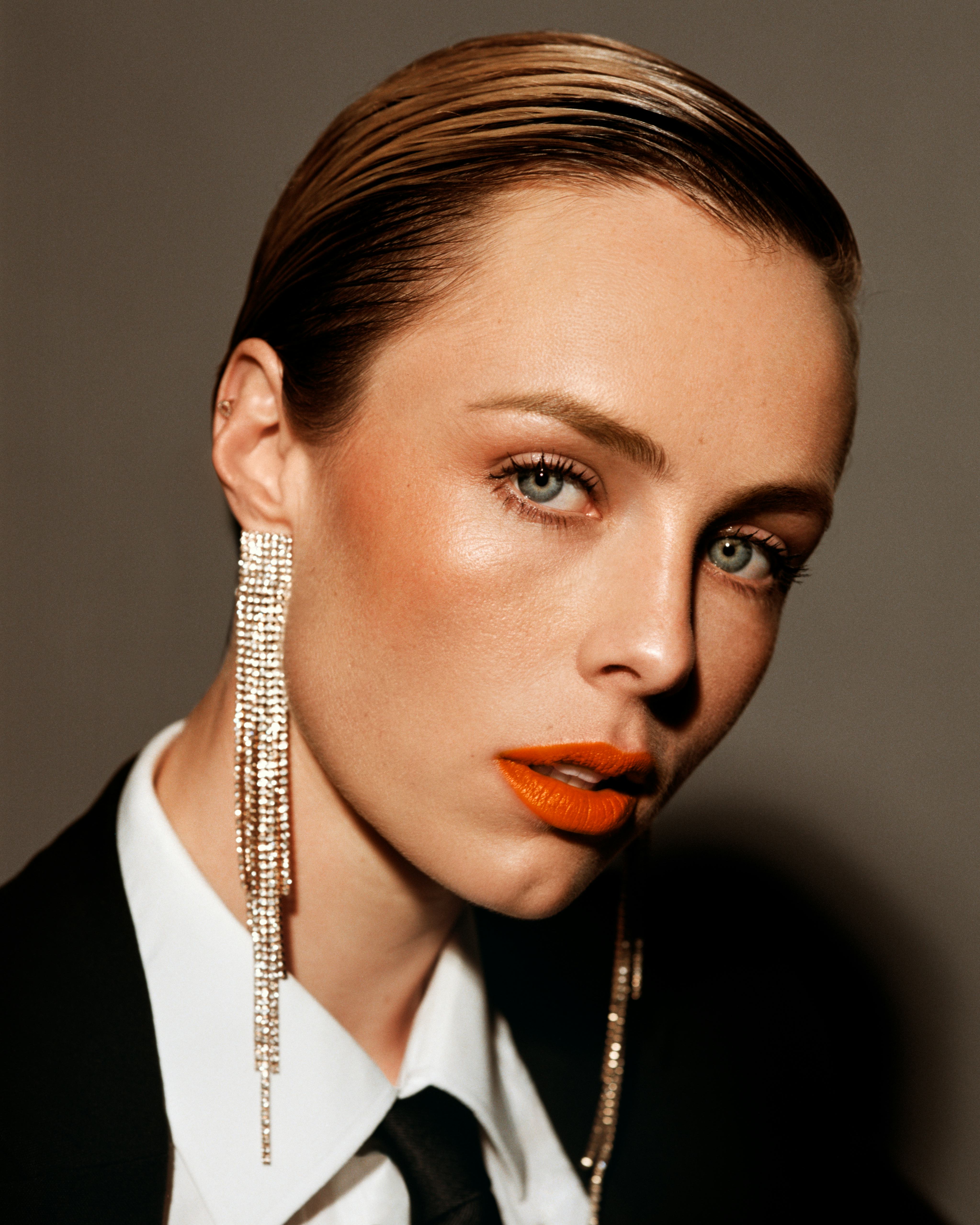 a person with an orange lip and chandelier earrings and slick back blonde bun