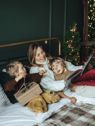 A mother and two children relax on a bed with a dog, surrounded by holiday decorations and Christmas...