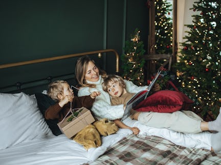 A mother and two children relax on a bed with a dog, surrounded by holiday decorations and Christmas...