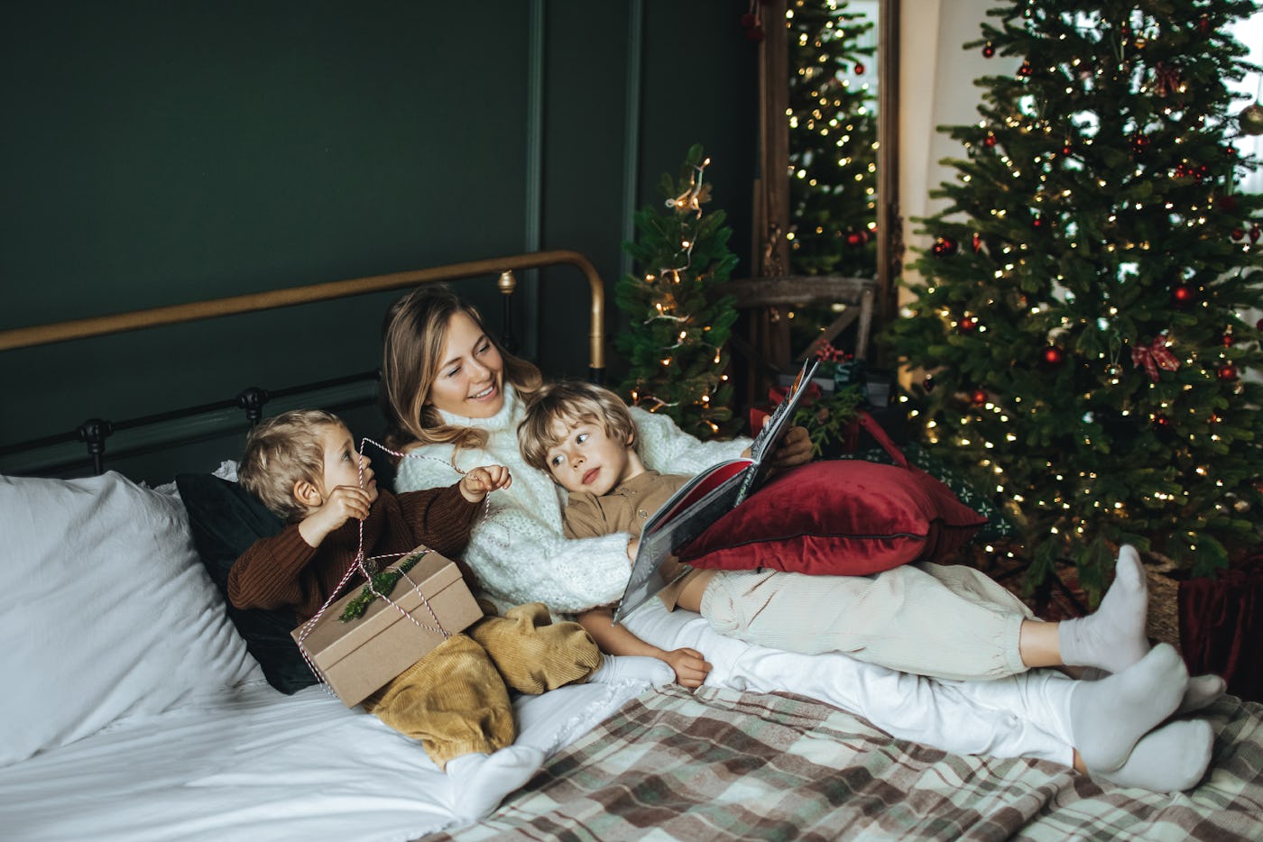A mother and two children relax on a bed with a dog, surrounded by holiday decorations and Christmas trees. One child holds a gift, while the other reads a book.