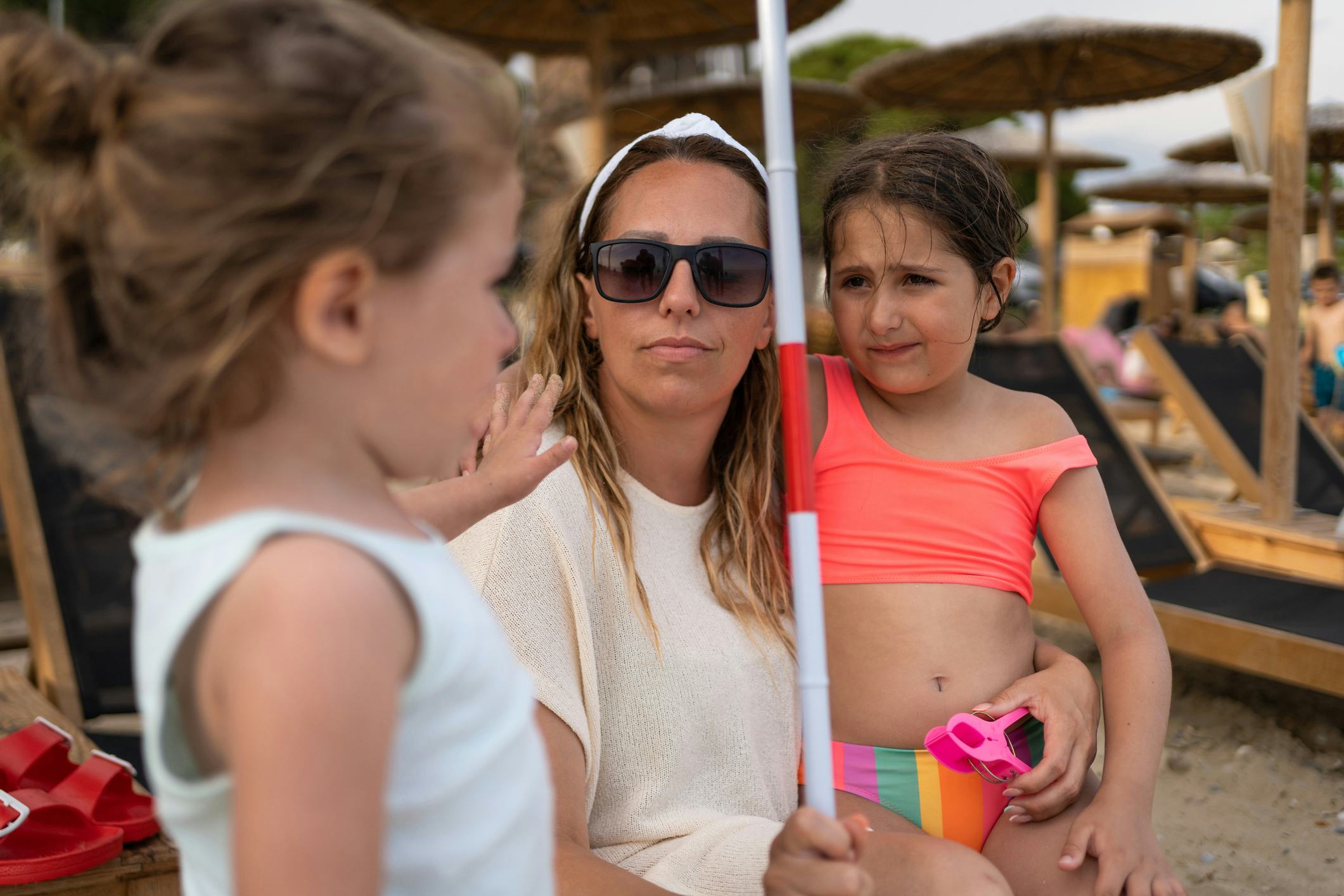 A woman in sunglasses holds a child wearing a pink swimsuit, while another child stands nearby, at a...