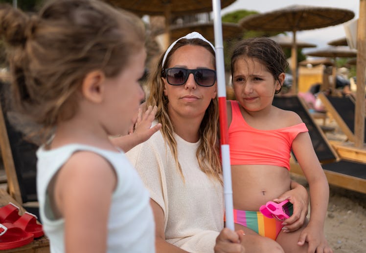 A woman in sunglasses holds a child wearing a pink swimsuit, while another child stands nearby, at a...
