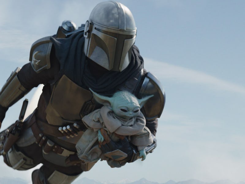 A Mandalorian warrior in sleek armor is flying through a desert landscape, securely holding a small,...