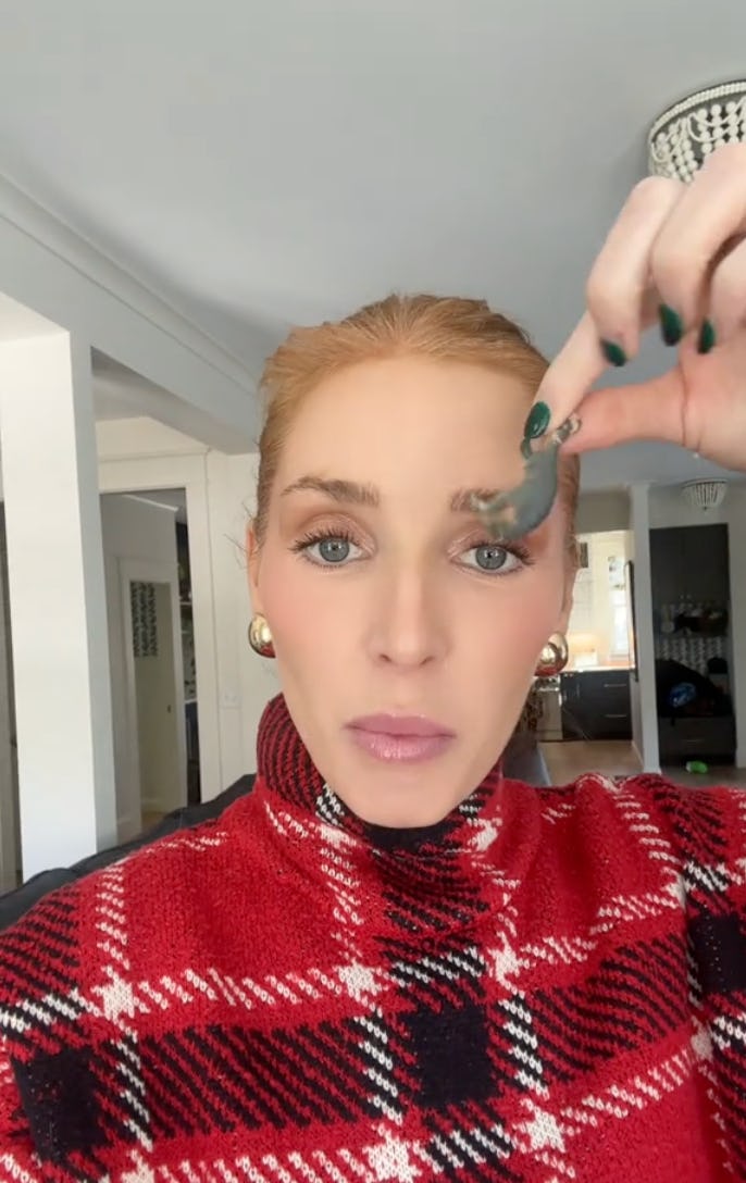 A woman in a red plaid sweater holds up a small toy. She has long hair, minimal makeup, and is in a bright indoor setting.