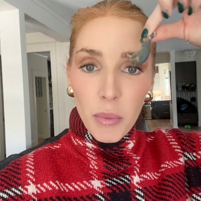 A woman in a red plaid sweater holds up a small toy. She has long hair, minimal makeup, and is in a bright indoor setting.