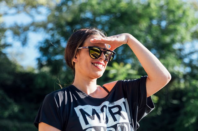 A smiling woman in a black T-shirt and sunglasses stands outdoors, shielding her eyes from the sun w...