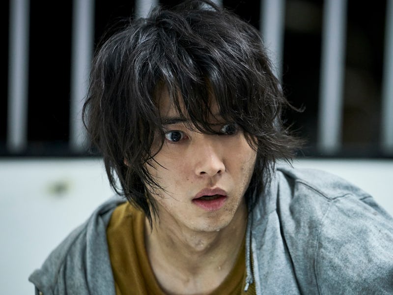 A young man with disheveled black hair looks surprised and anxious, wearing a gray hoodie over a yel...