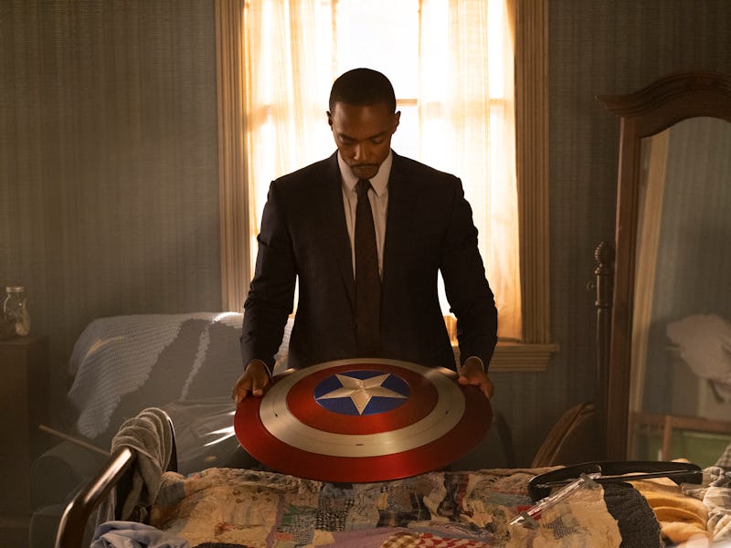 A man in a suit stands by a bed, holding a large shield with a star and red, white, and blue colors,...