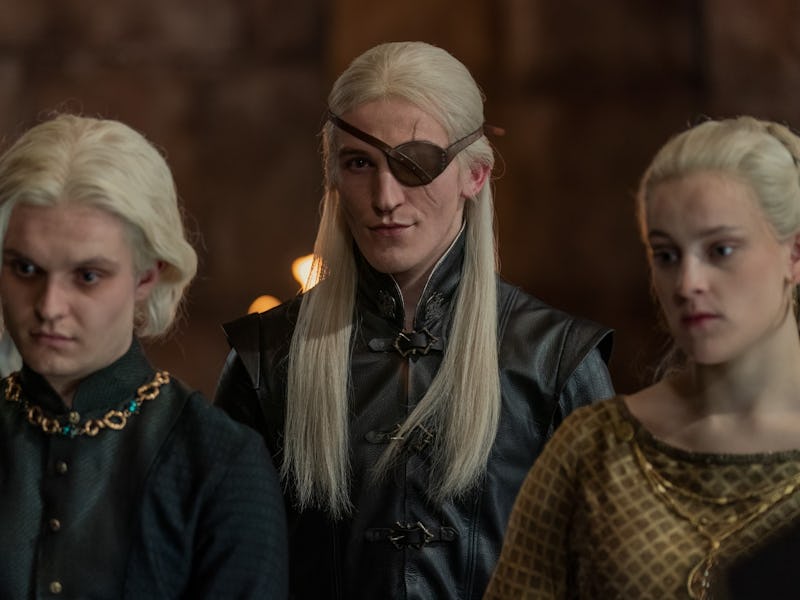 Three individuals with long, light-colored hair stand in a dimly lit room. One wears an eye patch an...