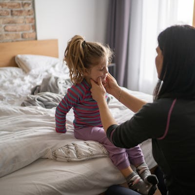A woman gently holds the cheeks of a young girl sitting on a bed, surrounded by soft bedding and natural light from a window.