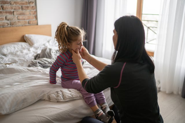 A woman gently holds the cheeks of a young girl sitting on a bed, surrounded by soft bedding and nat...