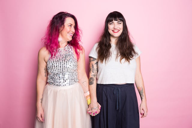 Two women stand side by side against a pink background, smiling and holding hands. One wears a sequi...