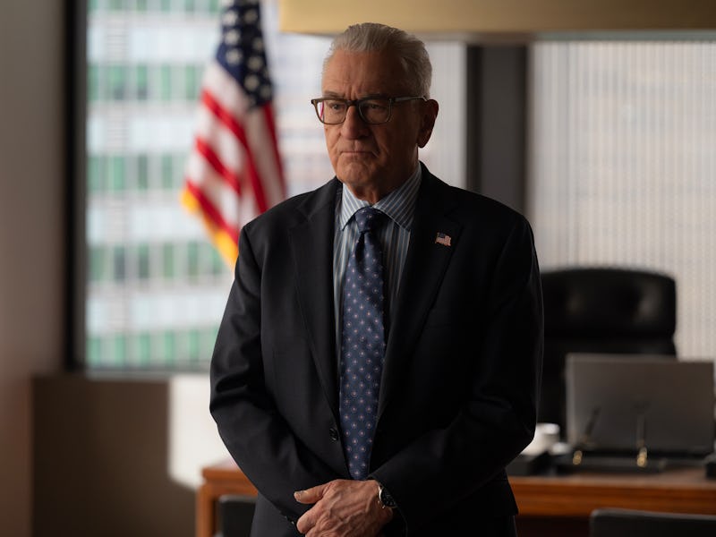 A distinguished older man with gray hair, wearing glasses and a suit, stands solemnly in an office w...