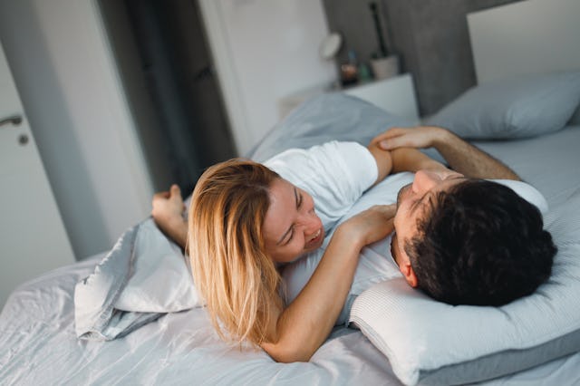 A couple lies on a bed, smiling and playfully engaging with each other. Soft bedding and a relaxed a...