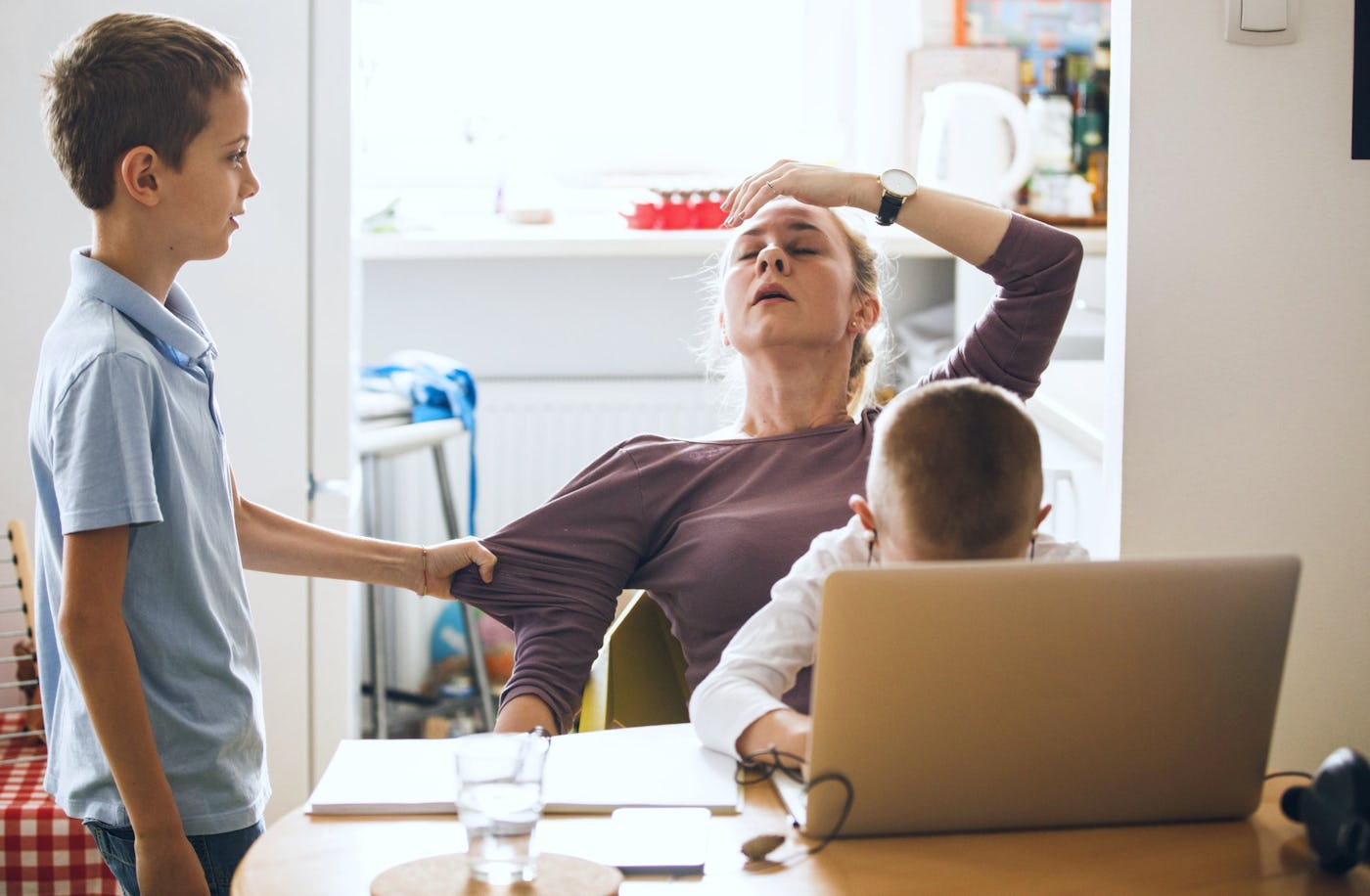 A woman looks tired at a table, while a boy stands beside her, touching her arm. Another boy with his head down sits at a laptop.