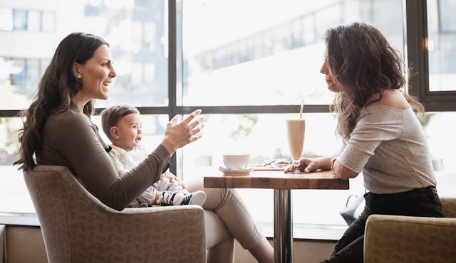 Two women are seated at a table in a cafe, engaged in conversation. One woman holds a baby, while th...