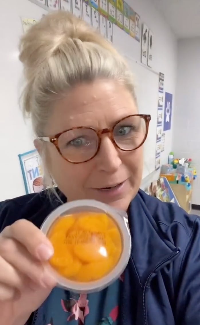 A smiling woman with a bun hairstyle holds a container of orange snacks, standing in a brightly lit classroom with colorful learning materials in the background.