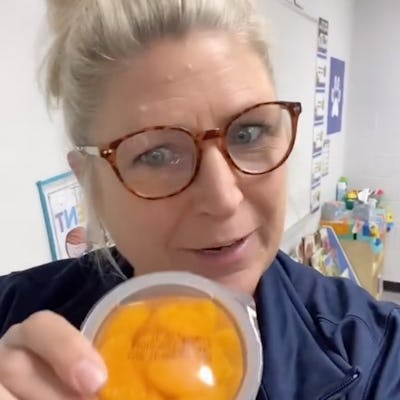 A smiling woman with a bun hairstyle holds a container of orange snacks, standing in a brightly lit classroom with colorful learning materials in the background.