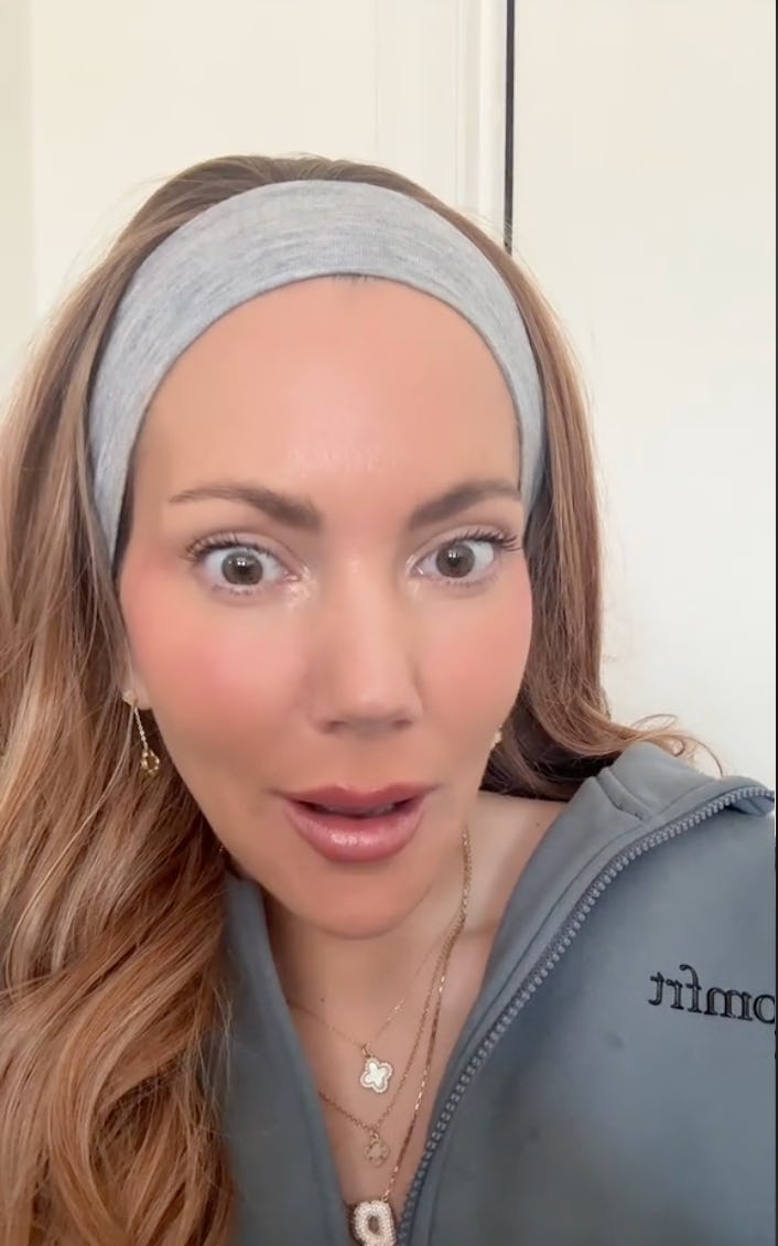 A woman with long, wavy hair and a gray headband looks surprised, wearing a casual gray outfit and delicate jewelry, set against a light background.
