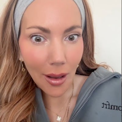 A woman with long, wavy hair and a gray headband looks surprised, wearing a casual gray outfit and delicate jewelry, set against a light background.