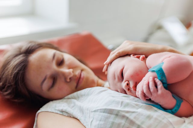 A mother rests peacefully with her newborn on her chest, sharing a tender moment in a softly lit roo...