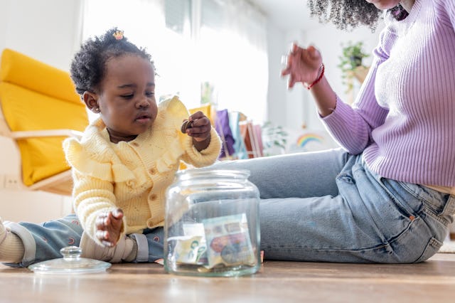 A young child in a yellow sweater is playing with coins while sitting on the floor next to an adult,...