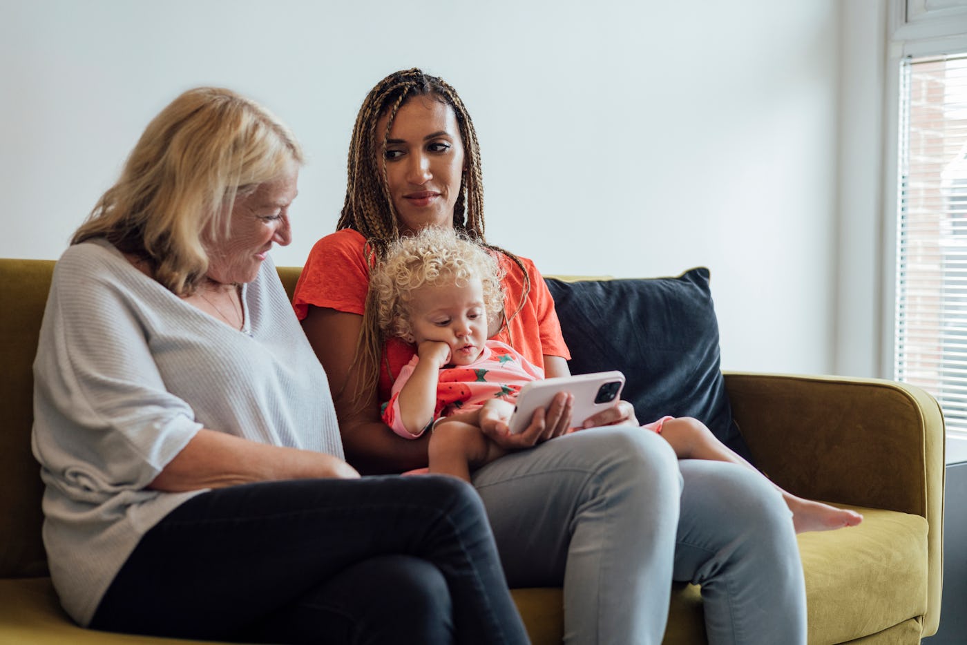 A woman with braided hair sits on a couch with a child and an older woman. They are engaged, looking at a phone together in a cozy living room.