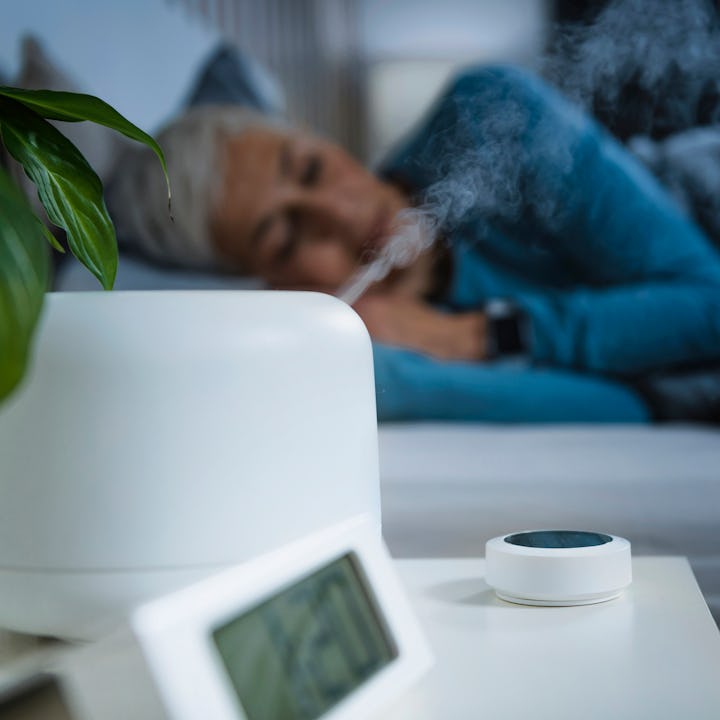 A cozy bedroom scene featuring a woman resting in bed while steam rises from a white humidifier. A p...
